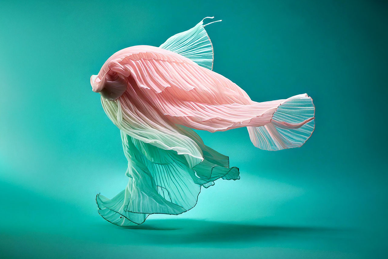 Surreal art photograph of a human figure draped in flowing pleated fabric resembling a betta fish, set against a teal background.