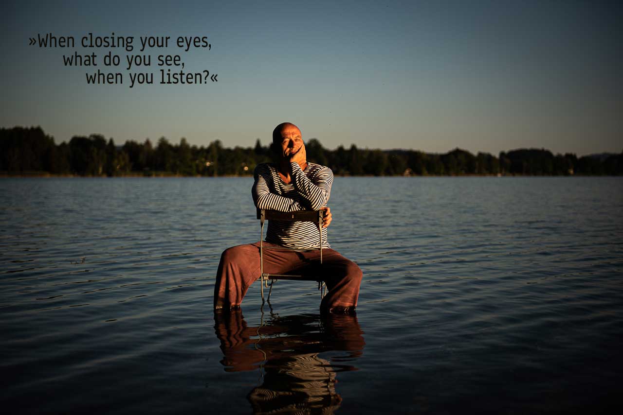 Maggowitsch sits on a chair partially submerged in water, his eyes closed, resting his head on one hand. The water reflects him as the golden sunlight casts long shadows. A poetic quote overlays the image: "When closing your eyes, what do you see, what do you listen."