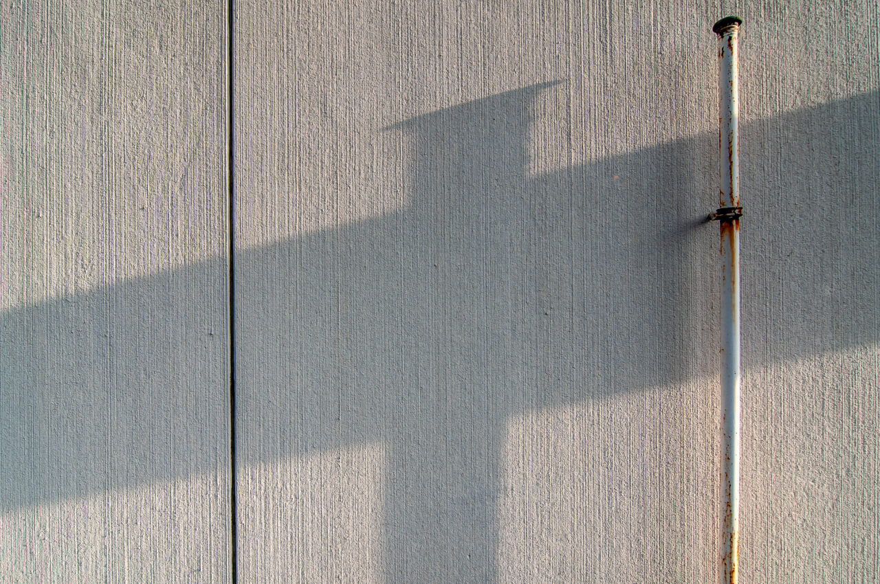 Schatten eines Rohrkreuzes auf einer Wand – symbolisiert stille Präsenz, Trennung und Durchlässigkeit zugleich.