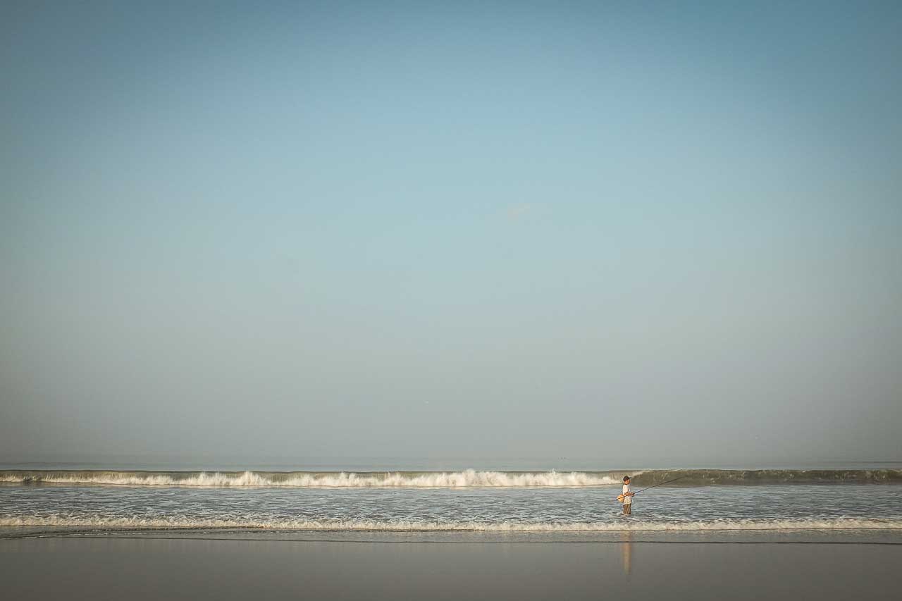 Ein man steht mit einer Angelk im flachen Wasser am Meer, vor ihm eine weite, stille Wellenlinie und ein klarer Himmel.