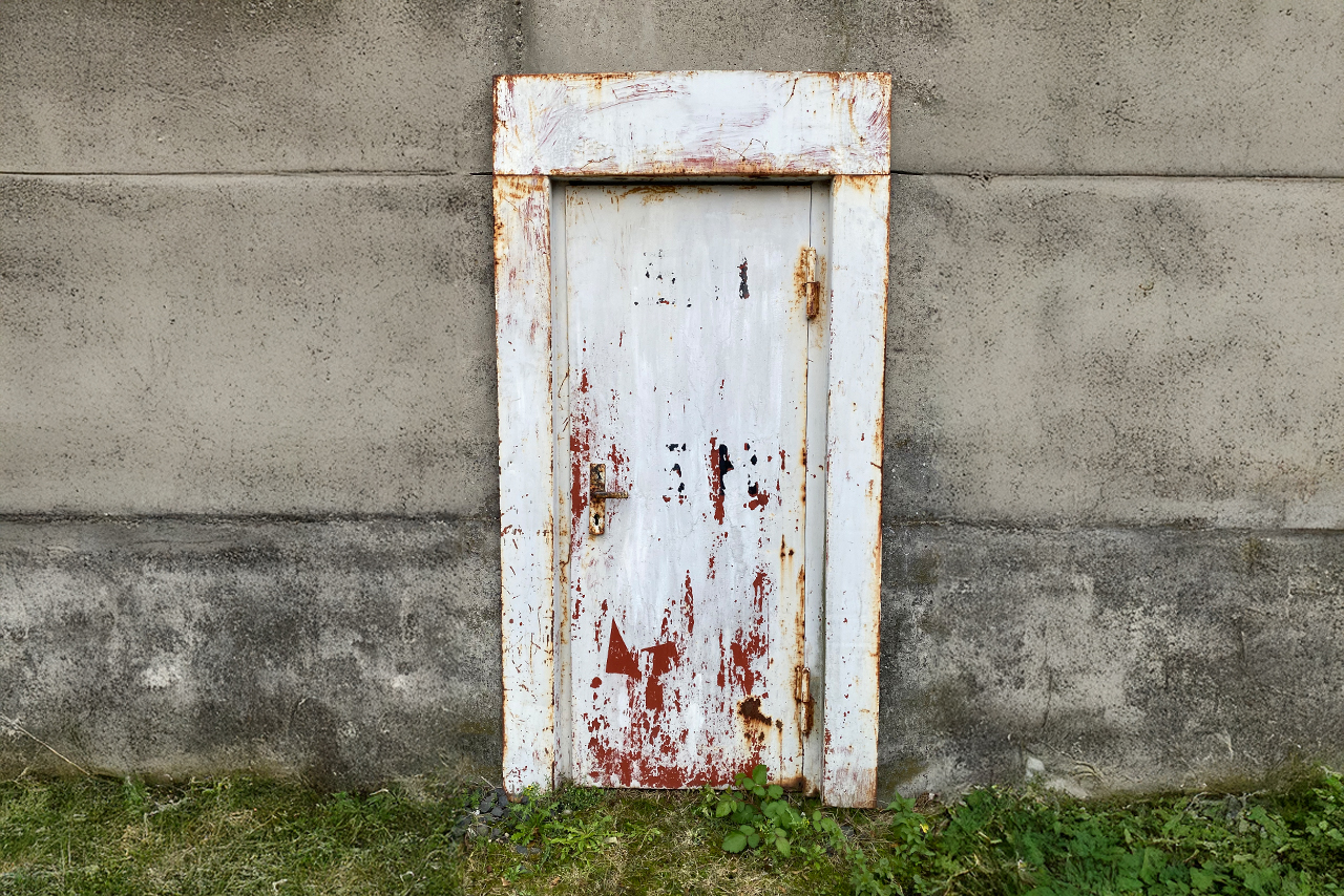 niedrige weiße tür in grauer wand, rostnarben und gras am sockel, stille, zurückhaltend, reduziert